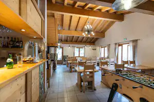 Innenraum mit Holzbar, Gläsern und Kicker-Tisch, viele Holztische und Stühle unter Lampen im Gemeinschaftsraum.