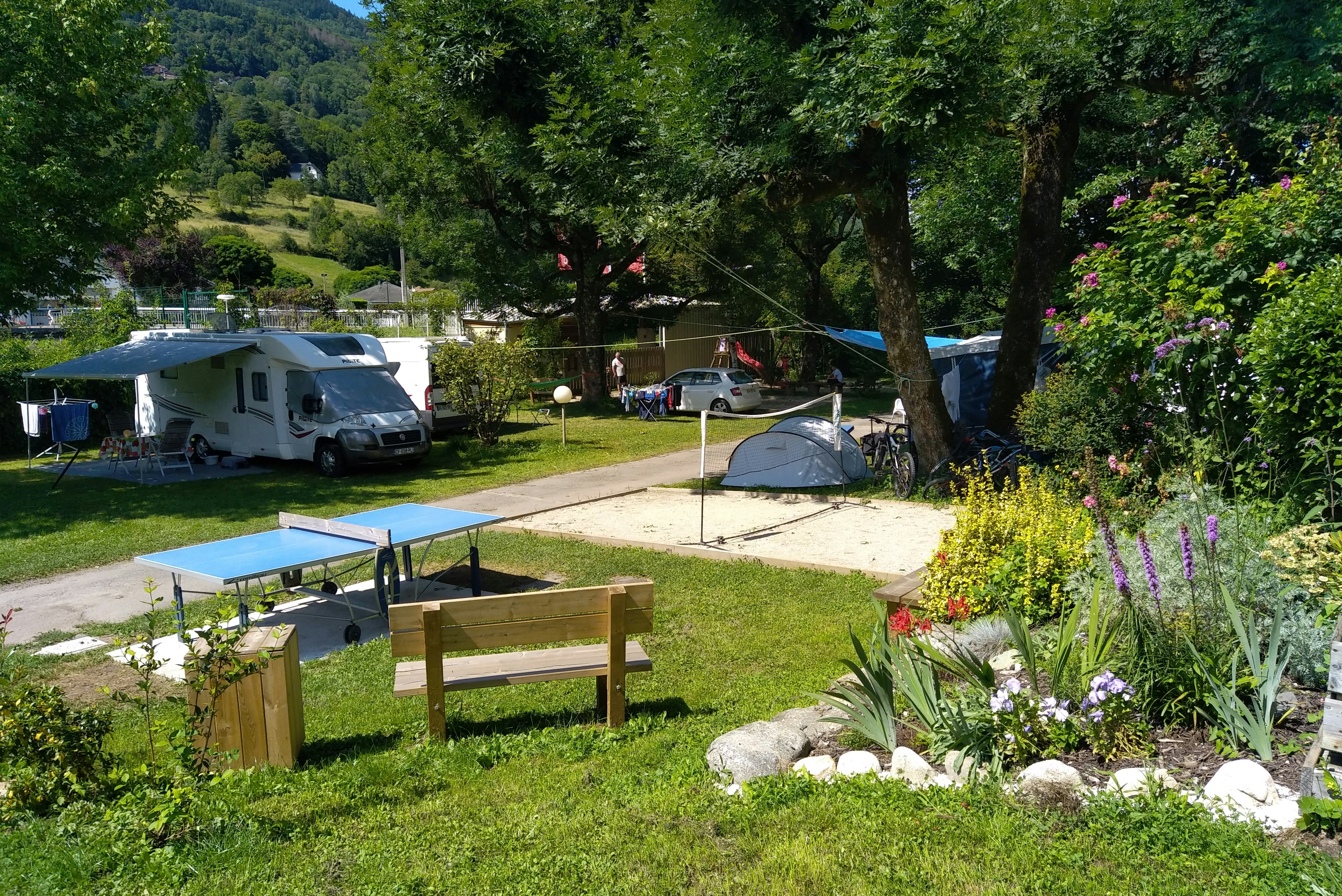 Wohnmobil und Zelt auf einem Campingplatz im Grünen, mit Tischtennisplatte, Gartenbank, Bäumen und Blumenbeeten.