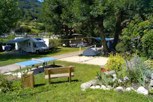 Wohnmobil und Zelt auf einem Campingplatz im Grünen, mit Tischtennisplatte, Gartenbank, Bäumen und Blumenbeeten.