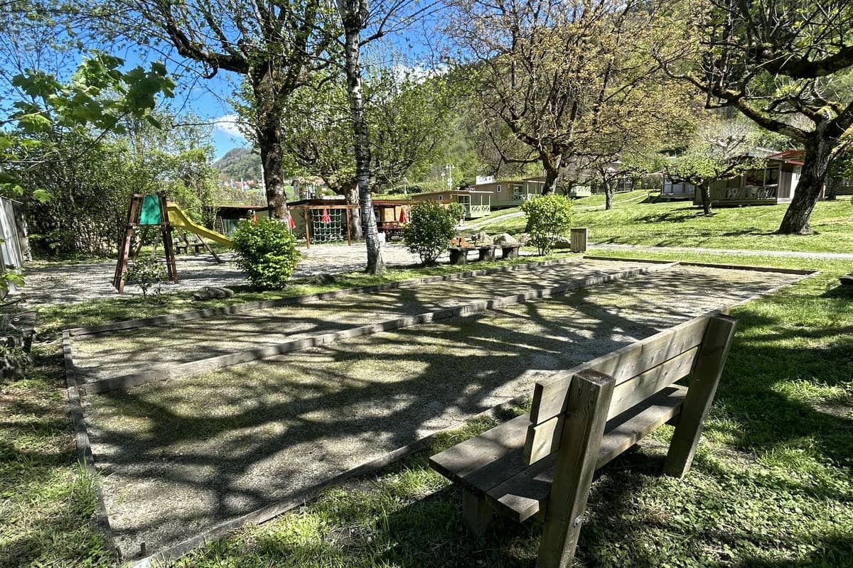 Holzbank und Terrasse auf einem Campingplatz zwischen Bäumen, mit Spielgeräten, Hecken und Wohnmobilen im Hintergrund.