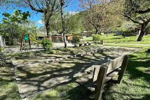 Holzbank und Terrasse auf einem Campingplatz zwischen Bäumen, mit Spielgeräten, Hecken und Wohnmobilen im Hintergrund.
