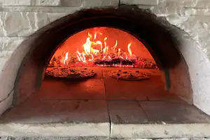 Flammen im Steinofen mit Feuerstelle, Ziegelboden und zwei Pizzablechen im Vordergrund.