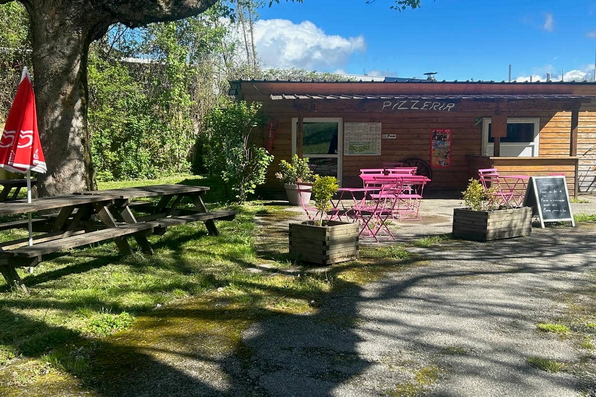 Terrasse einer Pizzeria am Campingplatz mit pinken Stühlen, Holzbänken, Pflanzkübeln und Weg vor dem Gebäude.