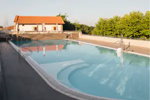 Schwimmbecken auf dem Campingplatz mit Sonnenterrasse, Blick auf das Poolhaus und Bäume im Hintergrund.