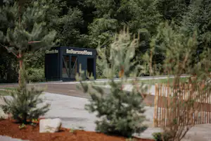 Informations- und Check-in-Gebäude am Campingplatz, umgeben von Bäumen und Hecken, mit Weg und Holzgeländer im Vordergrund.