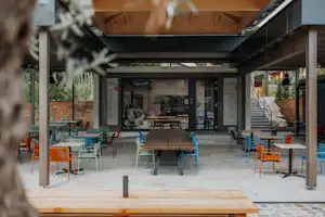 Überdachte Camping-Terrasse mit bunten Stühlen und Tischen vor einem kleinen Markt und großen Fenstern.