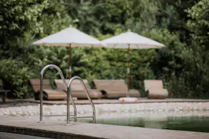 Camping-Pool mit Leiter, Holzdeck und zwei weißen Sonnenschirmen im Hintergrund, umgeben von Bäumen und Liegen.