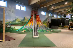 Große Kinder-Rutsche in einer überdachten Spielhalle mit buntem Rutschentuch, Wandbildern und grünen Matten.
