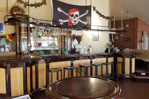 Barbereich im Campinghaus: Holztheke mit Biergläsern, Flaschenregal, Ziegelwand und Piratenbanner über der Theke.