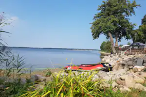 Roter Kajak am Ufer eines Sees, Steinkante und Gras vor einem Baum, im Hintergrund ruhiges Wasser.