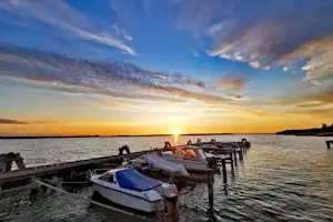 Bootssteg am See bei Sonnenuntergang, mehrere Boote liegen fest vertäut, im Hintergrund Wasser und Himmel mit Wolken.