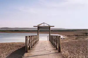 Holzsteg mit Seilgeländer führt über den Strand zum Wasser, im Hintergrund Küste und Hügel.