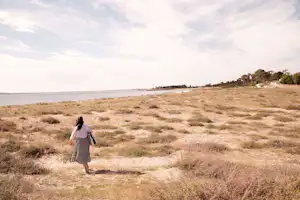 Frau mit Rucksack geht am Strand entlang, Blick auf das Meer, trockene Wiese und Bäume am Ufer.