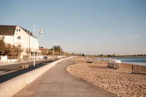 Promenade am See mit gepflastertem Weg, Sandstrand und weißen Gebäuden; Laternen und Zäune säumen die Uferlinie.
