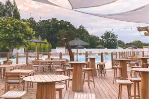 Holzterrasse mit vielen Stühlen und Tischen, Blick auf den Pool und Sonnenschirme zwischen Bäumen im Campingbereich.