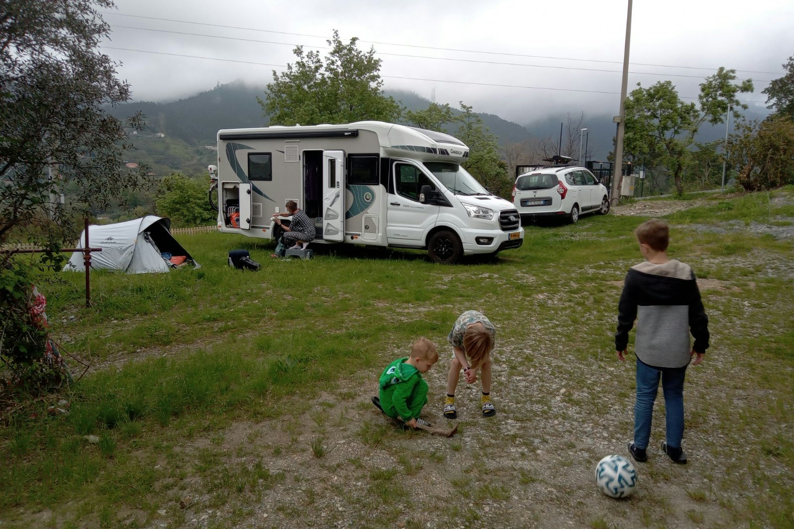 Wohnmobil auf dem Campingplatz mit Vorzelt, daneben Auto und Zelt, im Hintergrund Bäume und Berge im Nebel.