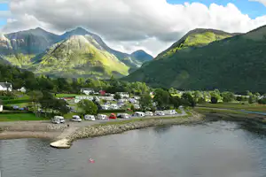 Campingplatz am See mit Wohnmobilen und Autos auf Stellplätzen, im Hintergrund grüne Berge und bewaldete Hänge.