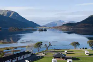 Campingplatz am See mit Wohnwagen, Rasenflächen und Blick auf Berge und ruhiges Wasser im Tal.