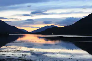 Abendstimmung am See mit spiegelnder Wasseroberfläche, Bergen im Hintergrund und weitem Himmel mit Wolken.