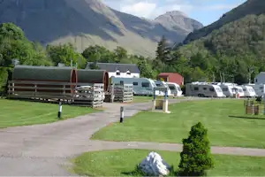 Campingplatz mit Wohnmobilen und Wohnwagen, gepflegener Rasenfläche und Kiesweg, im Hintergrund hohe Berge und Bäume.