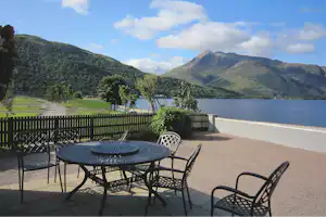 Terrasse mit Tisch und Stühlen am See, Blick auf Wasser, Bäume und bewaldete Berge im Hintergrund.