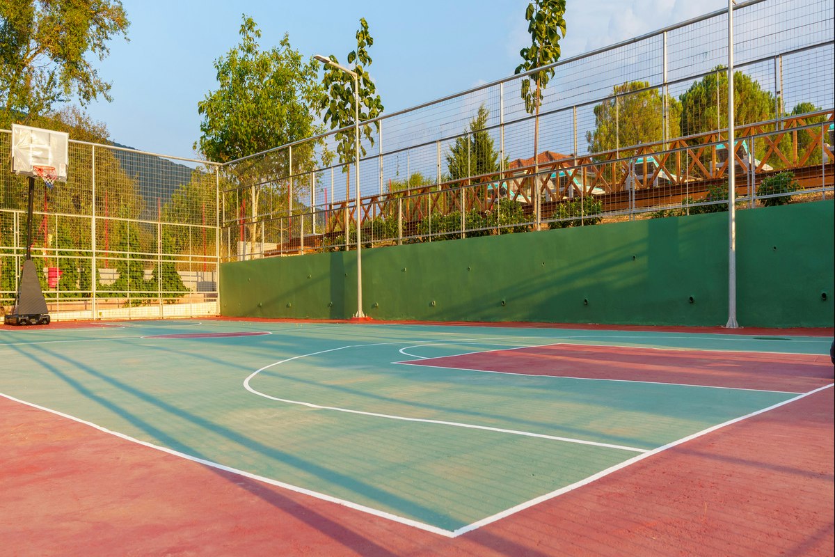 Sportplatz auf dem Campinggelände mit Basketballkorb, Kunstrasen und Umzäunung, dahinter Bäume und ein Gebäude.