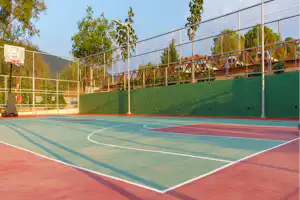 Sportplatz auf dem Campinggelände mit Basketballkorb, Kunstrasen und Umzäunung, dahinter Bäume und ein Gebäude.
