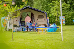 Familiencamping: Zelt mit Vorhang, Campingmöbel und Spielbereich mit blauem Spielgerät im grünen Garten zwischen Bäumen.