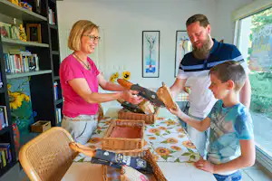 Familie am Esstisch in einer Ferienwohnung: Mutter und Vater öffnen Geschenke, Kind schaut zu, Bücherregal im Hintergrund.