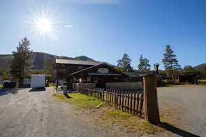 Holzgebäude mit Terrasse und Zaun am Campingplatz, Schotterweg, Bäume und Berge im Hintergrund, sonniger Himmel.