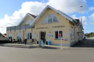 Camping-Rezeption und Turistbureau Gebäude mit großen Fenstern, Eingangstür und Sitzgruppe vor der Fassade.