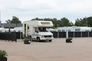 Wohnmobil auf einem Campingplatz-Stellplatz, mit Fahrrad am Eingang und Sicht auf weitere Fahrzeuge und Hecken.