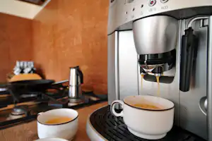 Kaffeemaschine mit zwei Tassen auf der Arbeitsplatte in einer modernen Küche, daneben Herd und Pfanne.