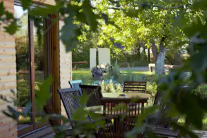 Terrasse am Campingplatz mit Holztisch und Gartenstühlen, Blick in den Garten mit Bäumen und Hecke.