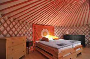 Holzbett mit gestreifter Matratze und Kissen in einem Zelt, daneben Kommode und Nachttisch, Holzdecke und rote Wände.
