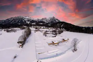 Winterlicher Campingplatz im Schnee mit Wegen, kleinen Holzhäusern und verschneiten Bergen im Hintergrund.
