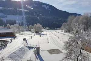 Winterlicher Campingplatz mit Stellplätzen, verschneiten Wegen und Zäunen, umgeben von verschneiten Wiesen und Bergen im Tal.