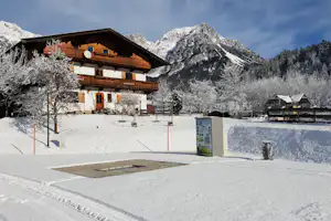 Alpen-Camping mit Holz-Chalet im Schnee, Bergkulisse im Hintergrund und gepflastertem Weg vor dem Gebäude.
