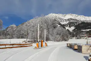 Verschneiter Campingplatz mit schmalem Weg, Holzstapeln und Stellplätzen, im Hintergrund hohe Berge und klare Winterlandschaft.