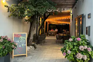 Camping-Terrasse mit Holzdach, Lichterkette und Sitzgarnitur, umgeben von Bäumen und Blumenbeeten vor dem Gebäude.