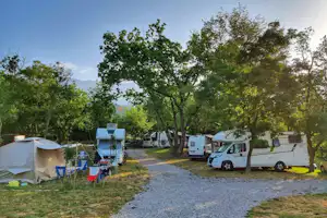 Zelt und Wohnmobile auf einem Campingplatz zwischen Bäumen, mit Kiesweg und Rasenfläche.