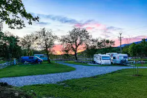 Wohnmobile und ein Auto auf einem Campingplatz, Kiesweg und Rasen zwischen Bäumen bei Sonnenuntergang.