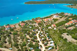 Luftaufnahme eines Campingplatzes am türkisfarbenen Meer mit Häusern, Wegen, Bäumen und mehreren Booten in der Bucht.