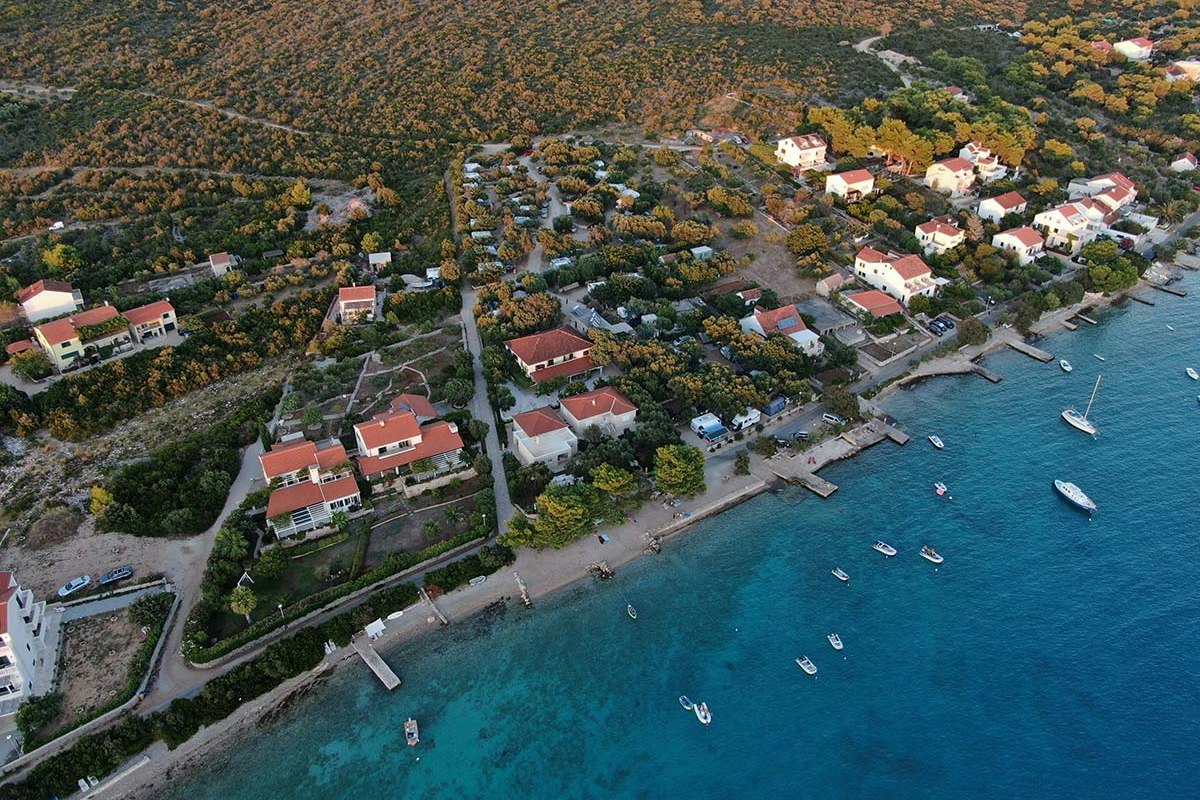 Luftaufnahme eines Campingplatzes am türkisfarbenen Meer mit Stegen, Booten und roten Dächern inmitten von Bäumen und Wegen.