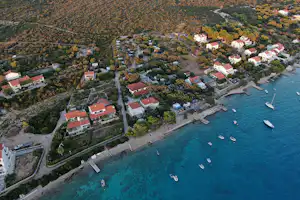 Luftaufnahme eines Campingplatzes am türkisfarbenen Meer mit Stegen, Booten und roten Dächern inmitten von Bäumen und Wegen.