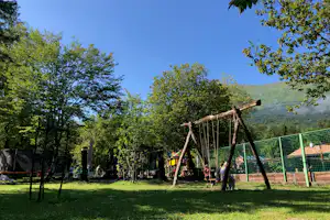 Spielplatz im Campingbereich mit Holzschaukel, grünen Zäunen und Bäumen vor grünen Hügeln im Hintergrund.
