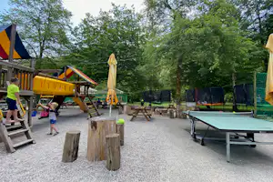 Spielplatz auf dem Campingplatz mit gelber Rutsche, Trampolin, Tischtennisplatte und Sitzbänken im Grünen.