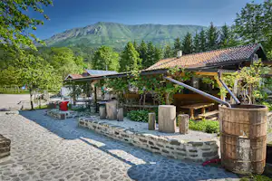 Holzterrasse mit Geländer, Sitzbank und Holzfässern am Campingplatz, im Hintergrund grünes Tal und Berge.