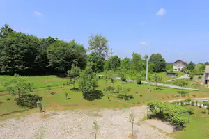 Weite Grünfläche eines Campinggeländes mit Bäumen, Wegen, Laternen und einem kleinen Teich im Hintergrund.