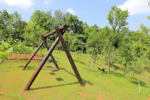 Schaukel aus Holz auf einer grünen Wiese, daneben Sitzbänke und Spielgeräte im Campinggelände.